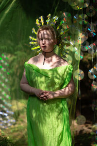 Young woman in mirrored mask and neon fabric standing in forest  front of the scenery from a cd case