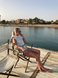 Young woman sitting on seat by lake against sky