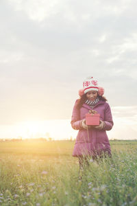 Woman with gift box standing on field against sky during sunset