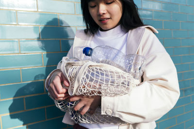 Young woman holding empty plastic bottles in mesh bag by turquoise brick wall