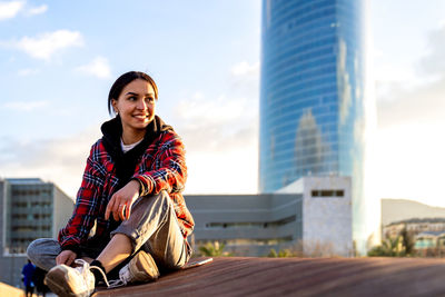 Portrait of smiling young woman sitting against sky in city