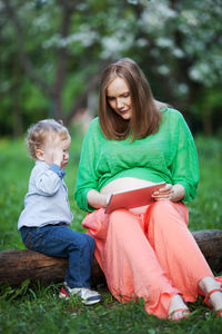 Pregnant mother showing digital tablet to son while sitting on log at public park