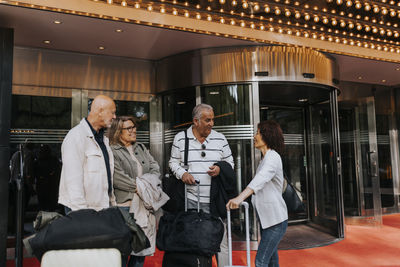 Male and female senior friends with luggage talking to each other while standing outside movie theater