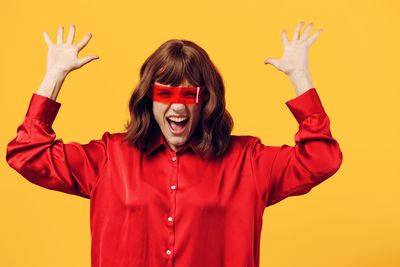 Portrait of young woman with arms raised standing against yellow background