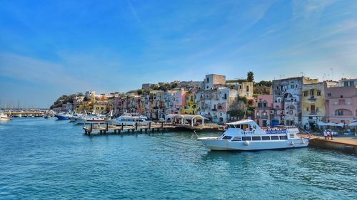 Sailboats moored on sea by buildings against blue sky