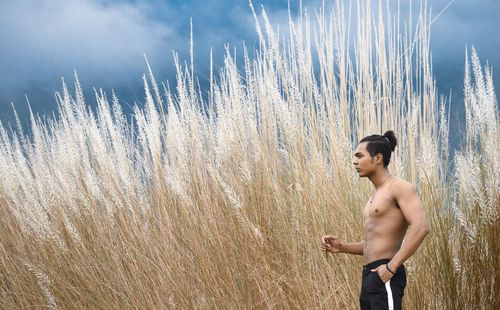 Side view of shirtless young man standing on field against cloudy sky