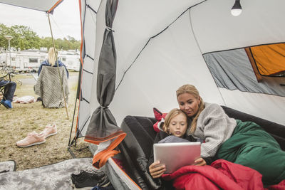 Girl watching digital tablet while lying with mother in tent at campsite