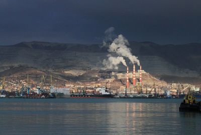 Commercial dock with ships and chimneys emitting smoke by mountain against sky