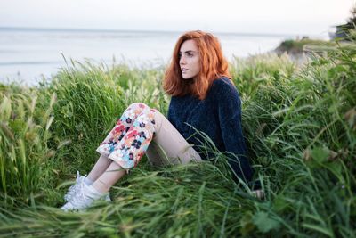 Woman sitting on grass by sea against sky