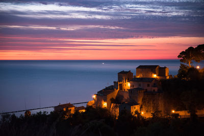 Illuminated building by sea against sky at sunset