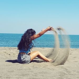 Woman playing with sand at beach against clear blue sky