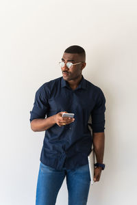 Serious african american adult male in stylish outfit and eyeglasses standing with netbook in hands while using cellphone and looking away on white background