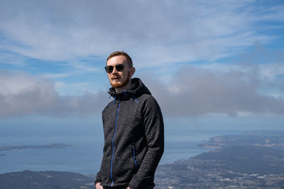 Young man wearing sunglasses standing on mountain against sky