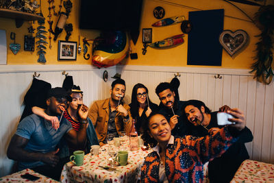 Young woman taking selfie with cheerful multi-ethnic friends holding props while sitting at restaurant during dinner par