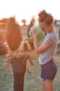 Side view of girl water plants while standing on grass against sky