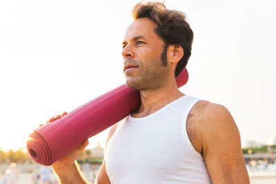 Calm middle aged male carrying rolled up mat on shoulder and looking away during yoga session at sunrise on beach