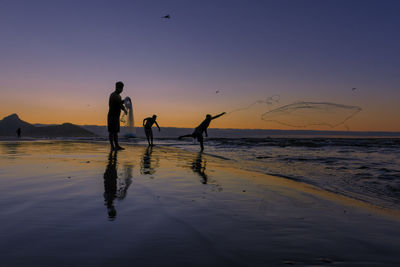 Silhouette people on beach throwing nets against sky during sunset