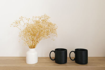 Dried flowers in a white ceramic vase and two black mugs on a wooden countertop in decor dining room