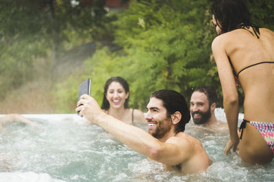 Young man taking selfie through mobile phone while friends enjoying in hot tub during weekend getaway