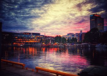 Illuminated buildings by river against sky at sunset
