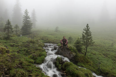 Man sitting on rock by stream at grassy field during foggy weather