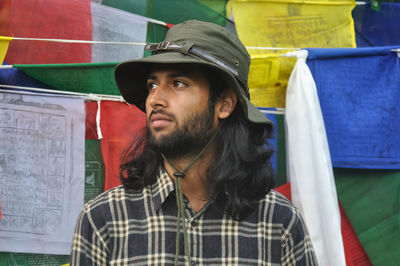 A bearded and long haired male tourist looking sideways while standing against tibetan prayer flags 