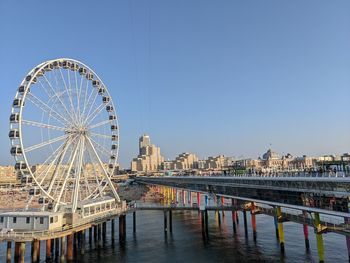 Ferris wheel by river and buildings against clear sky