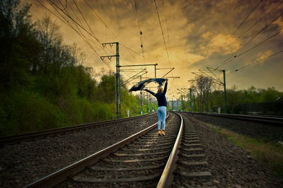 Rear view of woman with arms outstretched holding scarf while standing on railroad track during sunset