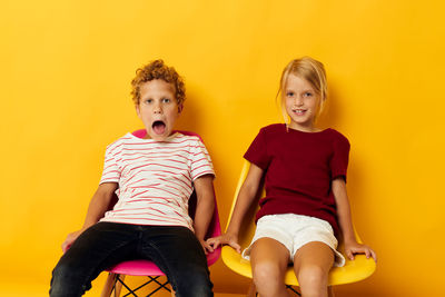 Portrait of smiling sibling sitting on chair against yellow background