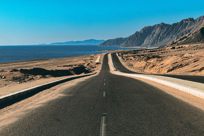 Empty road leading towards mountains against clear blue sky