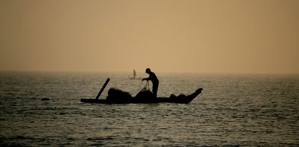 Silhouette fishermen in boats sailing over sea against clear sky during sunset