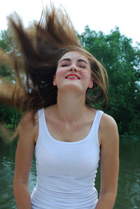 Cheerful young woman tossing hair while standing against lake in park