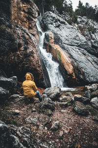 Rear view of man sitting on rock at waterfall