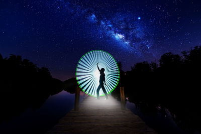 Digital composite image of man dancing against light painting on pier by lake at night