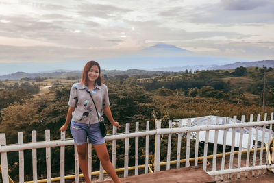 Portrait of smiling young woman standing on railing against sky