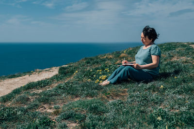 Adult woman thoughtfully writes plans diary on a sunny day on a mountain against backdrop of the sea