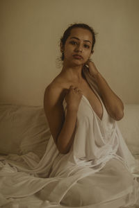 Portrait of shirtless young woman sitting on bed against wall at home
