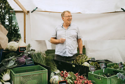 Happy senior male owner looking away while standing amidst vegetables crates at flea market