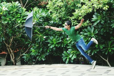 Man with umbrella jumping on walkway against trees