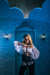 Portrait of woman in costume holding sword standing against door of abandoned building at night