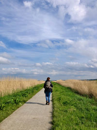 Rear view of woman walking on road amidst field against sky
