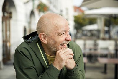 Elderly bald man with beard, laughing in a green marmot jacket, seated at an cafe during the day