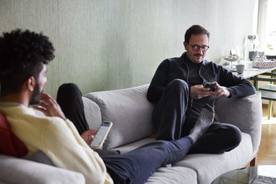 Gay couple relaxing on sofa at home and using phones
