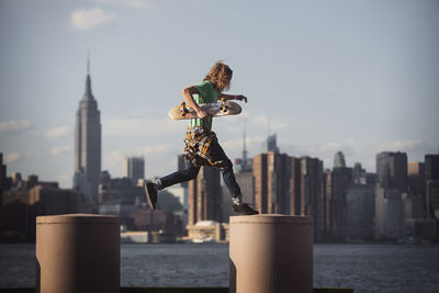 Man holding skateboard while jumping on columns by river in city