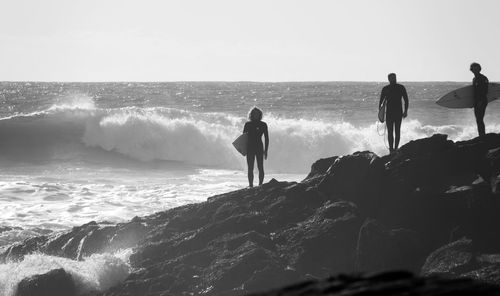 Surfers standing on rocks at beach against sky