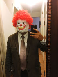 Reflection of man wearing clown mask while photographing with mobile phone on mirror