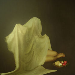 Naked girl hiding under fabric by fruits against black background