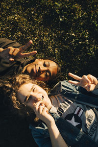 High angle portrait of smiling friends gesturing peace sign while lying on field during sunny day