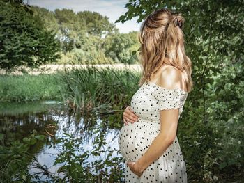 Side view of pregnant woman standing at lakeshore against trees