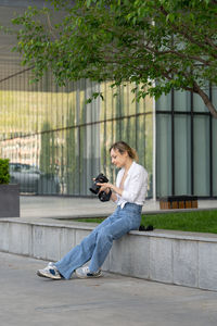 Focused young woman sitting on bench in spring park under tree outdoors resting using camera